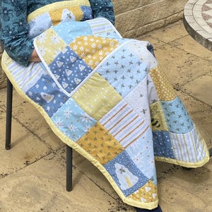 May include: A patchwork quilt with a yellow border, featuring squares of blue, white, and yellow fabric. The quilt has bee and floral patterns. The quilt is draped over a person sitting on a metal chair.
