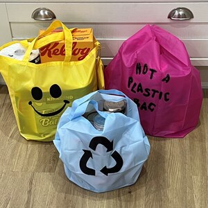 May include: Three reusable shopping bags. A yellow bag with a smiley face and the text "Kind and" and "Bath" visible. A pink bag with the text "Not a Plastic Bag". A blue bag with a recycling symbol.