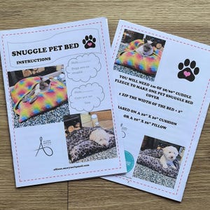 Puede incluir: Dos hojas de instrucciones para una cama Snuggle Pet. Las hojas presentan imágenes de gatos y perros en camas para mascotas. El texto incluye "Snuggle Pet Bed Instructions" y "Necesitará 1m de forro polar de 147/152cm para hacer una funda de cama para mascotas."