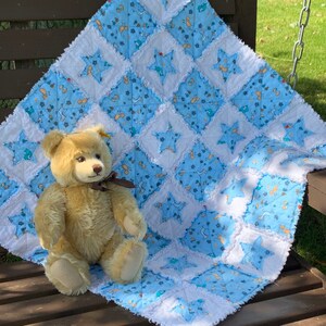 May include: A blue and white baby quilt with a star pattern and a teddy bear sitting on top of it.