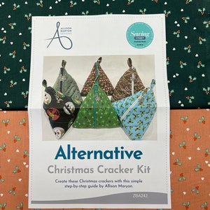 May include: A white paper pattern with a green and white holly pattern on the top and a red and white holly pattern on the bottom. The paper has a title that reads "Alternative Christmas Cracker Kit" and instructions to "Create these Christmas crackers with this simple step-by-step guide by Allison Maryon." The paper also has the text "ZBA242".