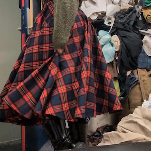 Può includere: Una gonna ampia e plissettata con un classico motivo a quadri rossi, blu navy e oro. La gonna è abbinata a stivali neri e un maglione verde. La gonna è in stile vintage, perfetta per un look retrò o preppy.
