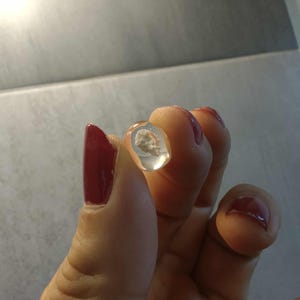 Könnte beinhalten: Ein klares, ovales Objekt mit einer kleinen, hellfarbigen Masse im Inneren. Das Objekt wird zwischen Fingern mit rotem Nagellack gehalten. Der Hintergrund ist ein hellgrauer Untergrund.