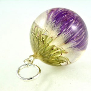 Puede incluir: Un colgante de esfera de vidrio transparente con una flor morada y blanca encapsulada en resina. El colgante tiene un bucle plateado para sujetarlo a una cadena o cordón.