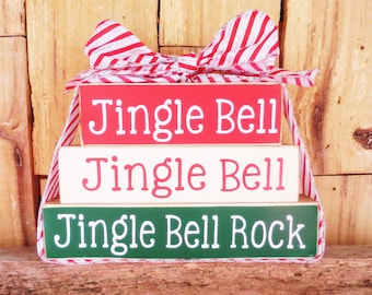 May include: A decorative Christmas sign with three stacked wooden blocks. The top block is red with "Jingle Bell" in white, the middle is white with "Jingle Bell", and the bottom is green with "Jingle Bell Rock". A red and white striped ribbon forms a bow at the top.