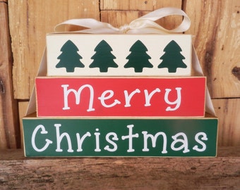 May include: A wooden Christmas sign with three tiers. The top tier is cream colored with four green Christmas trees. The middle tier is red with the word "Merry" in white. The bottom tier is green with the word "Christmas" in white.