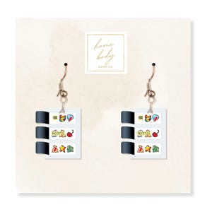 May include: A pair of silver-tone earrings featuring a slot machine design. Each earring has a rectangular shape with three windows displaying colorful icons like cherries, stars, and gems. The earrings are suspended from a fish hook.