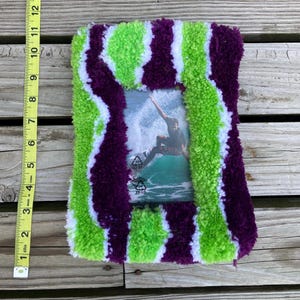 May include: A handmade photo frame with a vibrant, textured design. The frame is adorned with alternating stripes of green, purple, and white yarn. The frame measures approximately 12 inches tall and 8 inches wide, and holds a photo of a surfer.