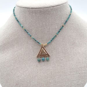 Op de afbeelding: Een gouden driehoek ketting met turquoise kralen. De hanger heeft een textuur en drie turquoise kralen hangen aan de onderkant.