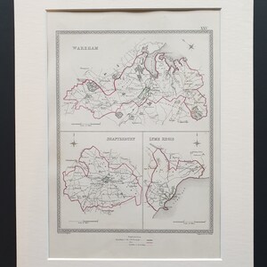 May include: An antique map print featuring three sections: Wareham, Shaftesbury, and Lyme Regis. The maps are detailed with roads, towns, and boundaries, framed in a white mat. The print has a vintage aesthetic.