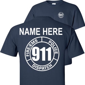 May include: Navy blue t-shirt with a white circle graphic on the back. The graphic features the text "FIRE/EMS POLICE DISPATCH" surrounding the number "911". The text "NAME HERE" is printed above the graphic.