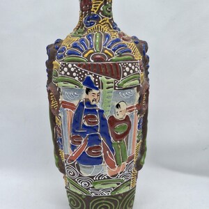May include: A decorative ceramic vase with a detailed, colorful design. The vase features a brown base and neck, with a central panel depicting figures in blue and red attire. The design includes floral and geometric patterns in green, yellow, and blue.