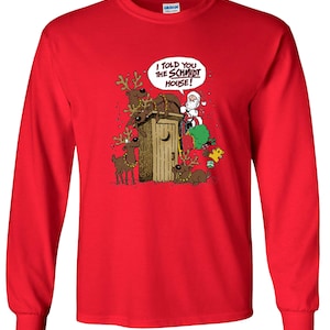 May include: A red long-sleeve shirt featuring a cartoon illustration of Santa Claus, reindeer, and a wooden outhouse. The text "I TOLD YOU THE SCHMIDT HOUSE!" is in a speech bubble. The shirt is made of a soft material.