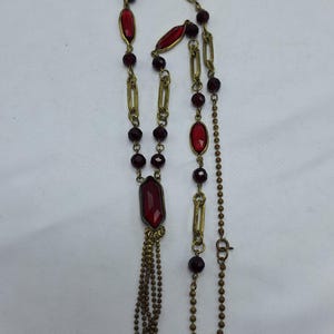 May include: A long necklace with red gemstone accents and a gold-tone chain. The necklace features oval and rectangular red stones, interspersed with small, round beads. A tassel of small chains hangs from the bottom.