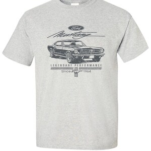 Peut inclure: T-shirt gris avec un motif vintage d'une voiture Ford Mustang noire. Le design comprend le logo Ford, le mot "Mustang" et le texte "LEGENDARY PERFORMANCE Since 1964". Le graphisme est vieilli pour un look rétro.