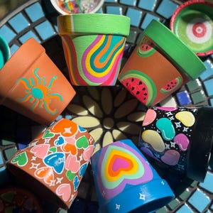 May include: A collection of six small terracotta plant pots, each hand-painted with distinct designs including colorful wavy lines, watermelon slices, a sun, hearts, and jelly beans, arranged in a circular pattern with matching saucers.