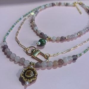May include: Three layered necklaces with beaded strands in shades of purple, white, and green. The top necklace has a gold clasp and a green bead. The bottom necklace has a flower pendant with a pearl.