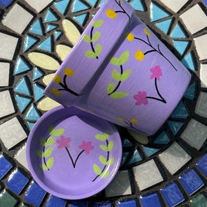 May include: A lavender-colored ceramic plant pot and matching saucer, both hand-painted with a pattern of thin black stems, light green leaves, small yellow circular buds, and pink four-petaled flowers scattered across the exterior surfaces.