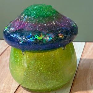 Op de afbeelding: Een paddenstoelvormige houder van hars met een fel limoengroene glitterbasis en een koepelvormige dop met een verloop van donkerblauw, paars en groen. De dop is ingelegd met holografische glitteraccenten in de vorm van sterren en maantjes.
