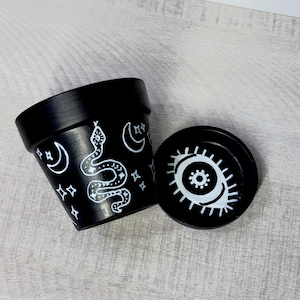 May include: A black terracotta plant pot and matching saucer. The pot features white hand-painted designs of a snake, crescent moons, and four-pointed stars. The saucer displays a white hand-painted eye motif with detailed eyelashes.