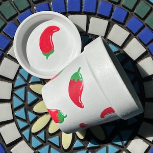 May include: A white ceramic plant pot and matching saucer, both decorated with hand-painted red chili peppers featuring green stems. The pot is positioned on its side, displaying the repeating chili pepper pattern across its exterior surface.