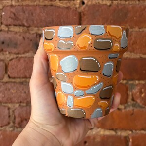 May include: A terracotta plant pot featuring a hand-painted pattern of smooth, rounded stones in shades of orange, brown, and light gray. The design covers the exterior surface of the pot, mimicking a stacked stone wall with small black dots.
