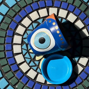 May include: A small terracotta plant pot painted in shades of blue, white, and black with a central eye motif and gold dot accents along the rim, accompanied by a matching solid bright blue circular saucer base for drainage and plant support.