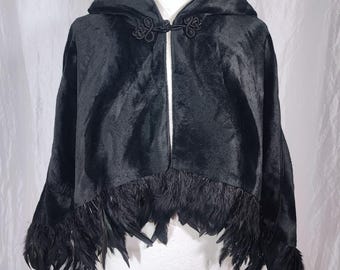 Op de afbeelding: Zwarte cape met capuchon en een verenrand. De cape heeft een decoratieve sluiting bij de halslijn. Het kledingstuk wordt op een mannequin getoond. De cape is gemaakt van een zacht, fluwelig materiaal.