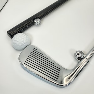 May include: A black and silver golf club license plate frame with a golf ball and a golf club.