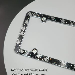 May include: A silver license plate frame with a black background and white paw prints. The frame is decorated with clear crystal rhinestones. "Genuine Swarovski Glass Cut Crystal Rhinestones" is written at the bottom of the image.