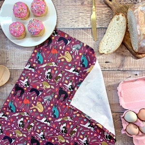 May include: A pink and white tea towel with a repeating pattern of black and white dogs, flowers, and leaves. The tea towel is folded in half and has a white backing.