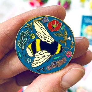 May include: A blue enamel pin featuring a yellow and black bumblebee surrounded by colourful flowers and leaves. The pin is circular and has a gold finish.
