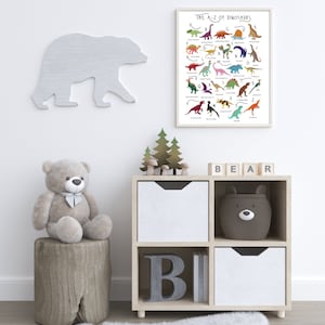 May include: A white wooden shelf unit with four compartments. The top two compartments have white drawers. The bottom left compartment has a large gray letter "B" and the bottom right compartment has a white drawer. A gray teddy bear sits on a wooden stump in front of the shelf. A white bear silhouette is on the wall behind the shelf. A framed poster with the text "The A-Z of Dinosaurs" is on the wall above the shelf. The poster features colorful illustrations of dinosaurs with their names listed in alphabetical order.