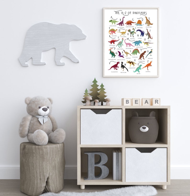 May include: A white wooden shelf unit with four compartments. The top two compartments have white drawers. The bottom left compartment has a large gray letter "B" and the bottom right compartment has a white drawer. A gray teddy bear sits on a wooden stump in front of the shelf. A white bear silhouette is on the wall behind the shelf. A framed poster with the text "The A-Z of Dinosaurs" is on the wall above the shelf. The poster features colorful illustrations of dinosaurs with their names listed in alphabetical order.