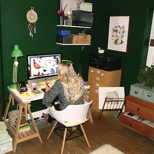 May include: A woman sits at a desk in a home office with a green wall, working on a computer. The desk is made of wood and has a white surface. There are several drawers and shelves in the room, as well as a window with blinds. The woman is wearing a black and white patterned shirt and has long blonde hair.
