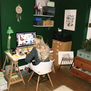 May include: A woman sits at a desk in a home office with a green wall, working on a computer. The desk is made of wood and has a white surface. There are several drawers and shelves in the room, as well as a window with blinds. The woman is wearing a black and white patterned shirt and has long blonde hair.