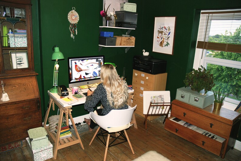 May include: A woman sits at a desk in a home office with a green wall, working on a computer. The desk is made of wood and has a white surface. There are several drawers and shelves in the room, as well as a window with blinds. The woman is wearing a black and white patterned shirt and has long blonde hair.