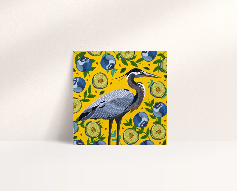 May include: A blue heron stands on a yellow background with a pattern of blueberries and lime slices. The heron has a long neck and beak, and its feathers are a mix of gray and blue.