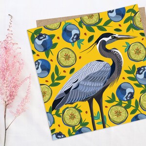 May include: A blue heron stands on a yellow background with a pattern of blueberries and lime slices. The heron has a long neck and beak, and its feathers are a mix of blue, gray, and white.