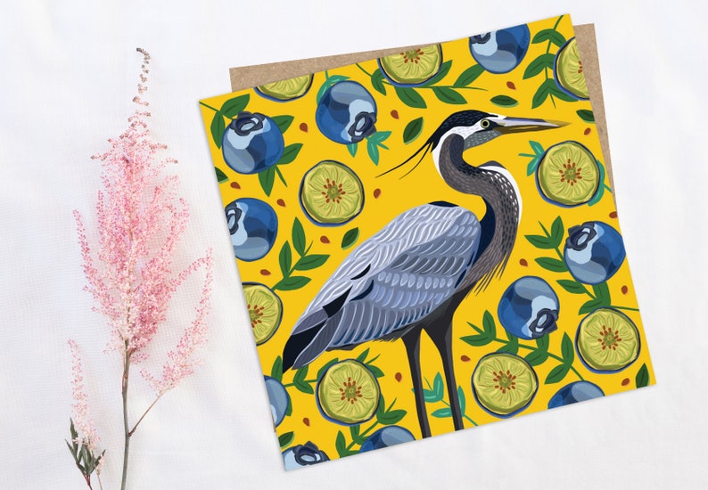 May include: A blue heron stands on a yellow background with a pattern of blueberries and lime slices. The heron has a long neck and beak, and its feathers are a mix of blue, gray, and white.