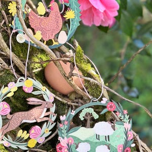 May include: Three colorful paper Easter ornaments hanging from a tree branch. The ornaments are shaped like circles and feature a floral wreath design. One ornament has a brown rabbit jumping in the center, another has a pink hen with chicks, and the third has a green field with white sheep.