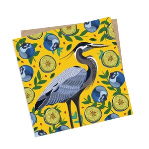 May include: A blue heron stands on a yellow background with a pattern of blueberries and green leaves. The heron has a long neck and a long beak. The blueberries are blue and the leaves are green.
