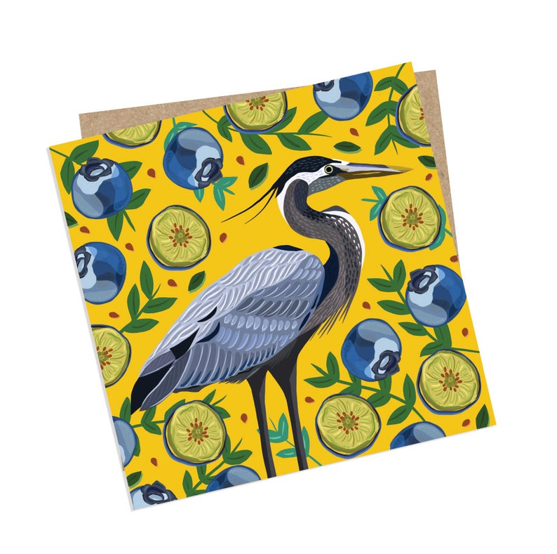 May include: A blue heron stands on a yellow background with a pattern of blueberries and green leaves. The heron has a long neck and a long beak. The blueberries are blue and the leaves are green.