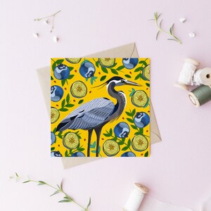 May include: A greeting card with a yellow background featuring a blue heron standing on a bed of green leaves and blue blueberries. The heron is surrounded by lemons and blueberries.
