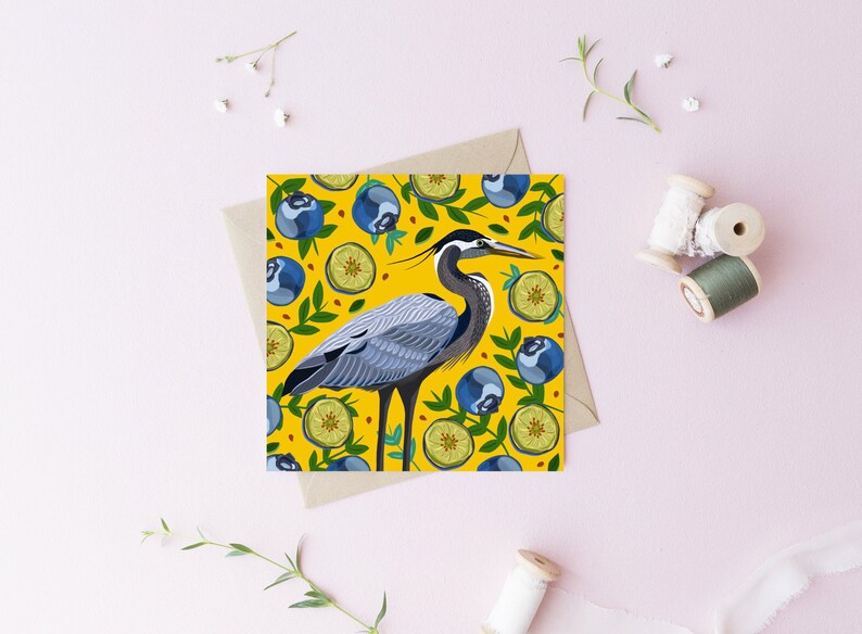 May include: A greeting card with a yellow background featuring a blue heron standing on a bed of green leaves and blue blueberries. The heron is surrounded by lemons and blueberries.