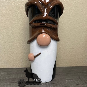 May include: A brown and white ceramic gnome mug with a black and brown face and a white beard. The mug has a brown pipe coming out of the side of the beard.