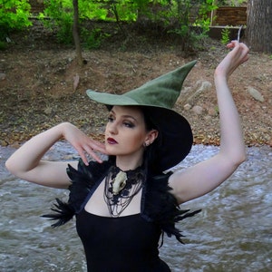 May include: A woman in a black dress and witch hat poses near a stream. She wears a feathered shoulder piece and a necklace with a skull pendant. The text "Queen Mab's" is visible at the bottom of the image.