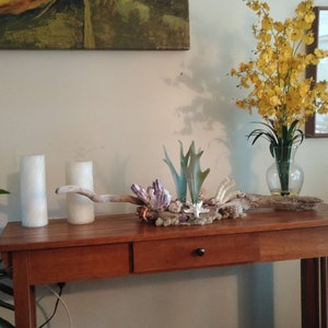 May include: A wooden table with two white candles, a driftwood centerpiece with seashells and a vase of yellow flowers.
