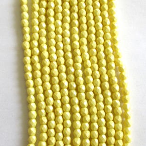 May include: Close-up of multiple strands of pale yellow faceted beads. The beads are uniform in size and shape, with a geometric design. The strands are arranged vertically against a white background, creating a textured pattern.
