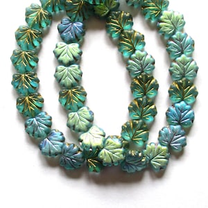May include: A strand of translucent glass beads shaped like leaves, featuring a gradient of teal and light green hues with gold-colored metallic accents highlighting the embossed vein details on the surface of each individual leaf-shaped bead.